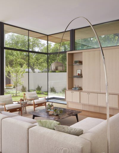 Modern living room with large windows, light wood furniture, and a curved floor lamp. A sofa faces two chairs and a wooden coffee table. Shelving and decor are in the background.