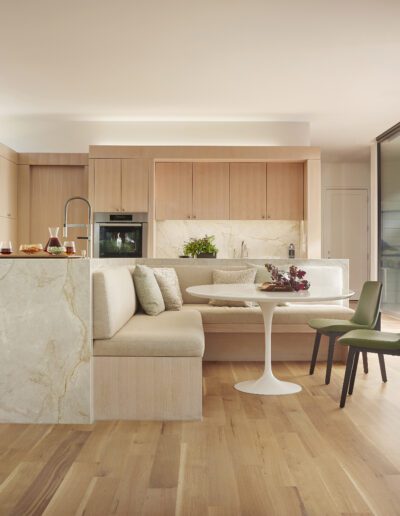 Modern kitchen with light wood cabinetry, a marble kitchen island, round dining table with green chairs, and an open living area with large windows.