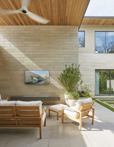 Modern patio with wooden furniture, beige stone walls, and large glass doors, partially covered by a wooden ceiling with a ceiling fan. Steps lead to a grassy section with tall plants.