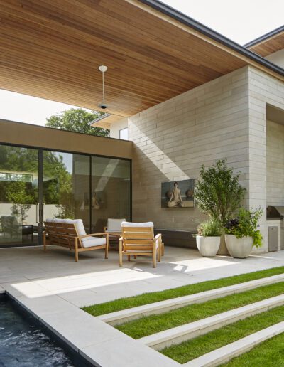Modern patio with wooden chairs, potted plants, and glass doors. Stone walls and ceiling overhang. Steps lead to grassy area and small pool.