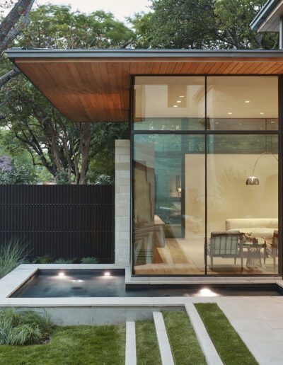 Modern house exterior with large glass windows, a small reflecting pool, and a landscaped yard with grass and trees.