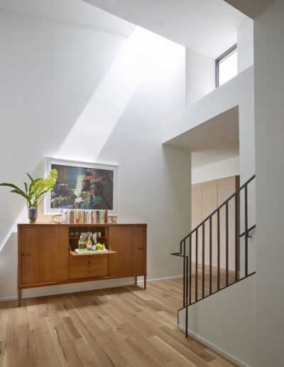 Modern interior with a wooden sideboard, a plant, books, art piece, and glassware. Light pours through a high window, illuminating the hardwood floor and open staircase with a black railing.