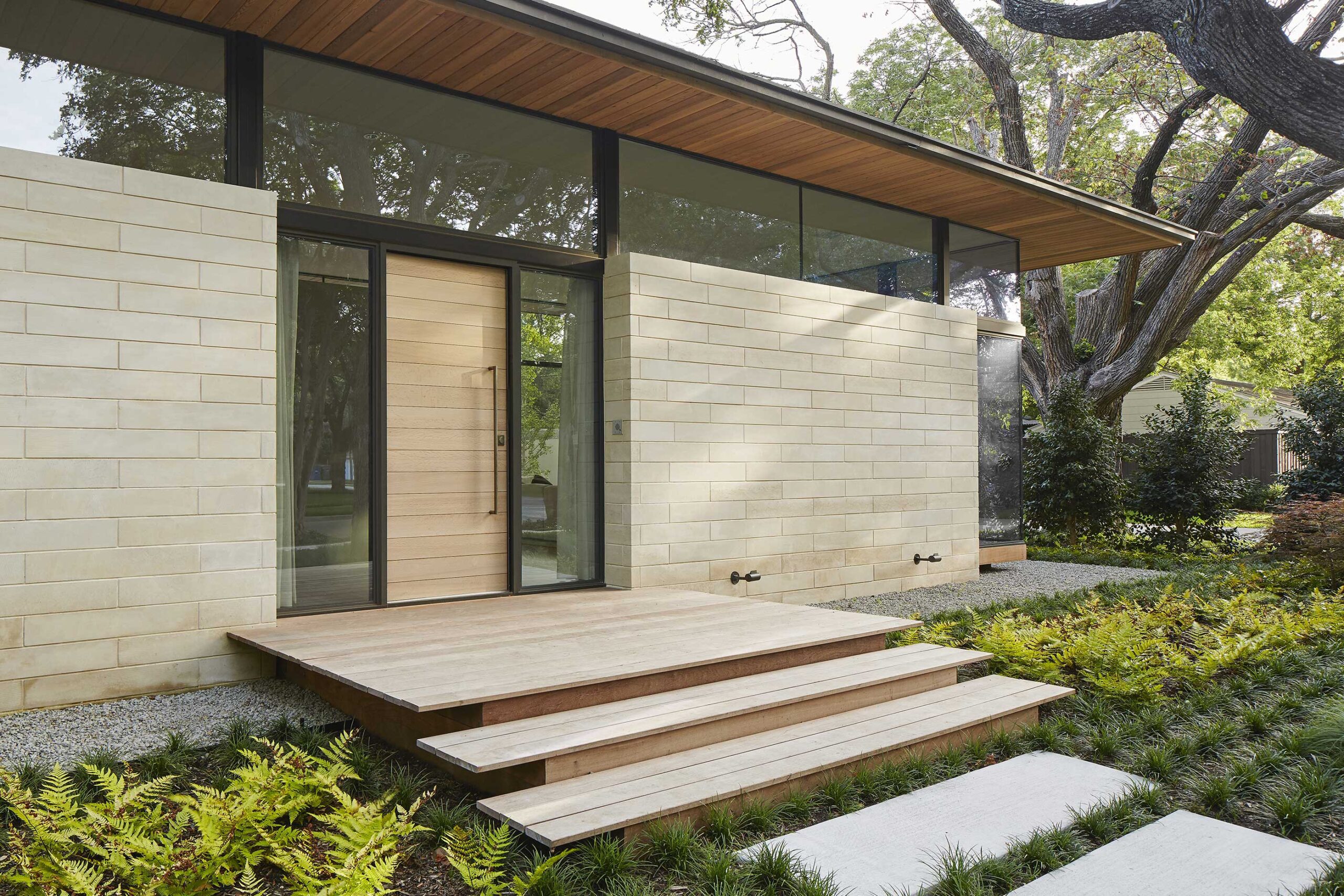 Modern house entrance with large glass door, light-colored stone walls, and wooden steps. Surrounded by greenery and trees.