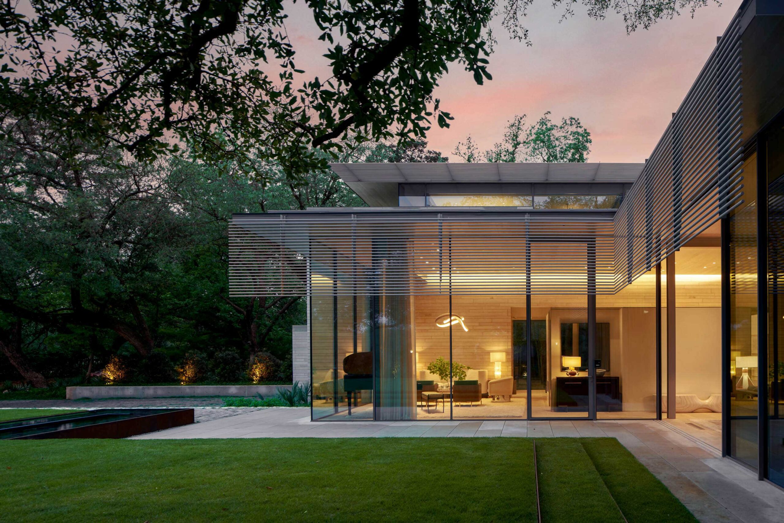 Modern house with large glass windows, flat roof, and a minimalist design. Surrounded by trees, with a manicured lawn in the foreground. Evening light casts a warm glow.