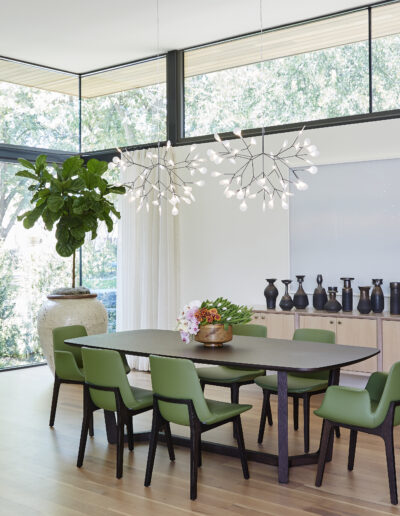 Modern dining room with a dark wood table, six green chairs, large window, hanging white light fixtures, potted plant, vase decor, and wooden sideboard against a white wall.