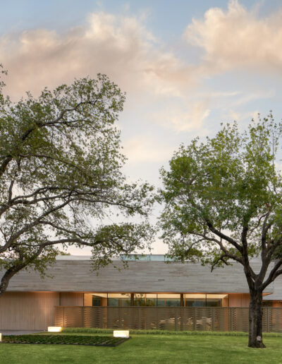 Modern house with a flat roof, large windows, and light-colored exterior, surrounded by trees and green lawn, under a partly cloudy sky.