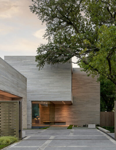 Modern house exterior with large concrete blocks, glass entrance, and a flat roof. A tree with green leaves partially frames the structure. Driveway with patterned gates in the foreground.