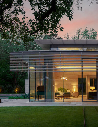Modern glass house exterior at dusk, surrounded by trees, featuring large windows and illuminated interiors.