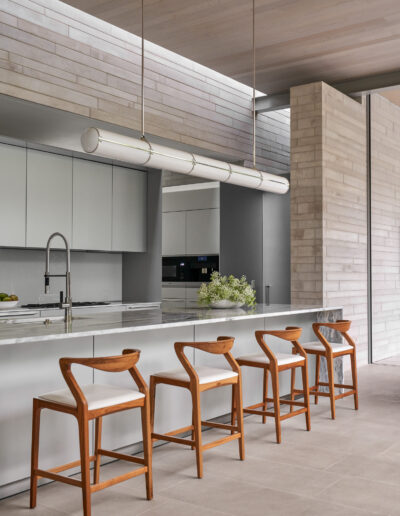 Modern kitchen with a marble island, four wooden stools, and a sleek cylindrical pendant light. Minimalist design with white cabinets and gray walls.