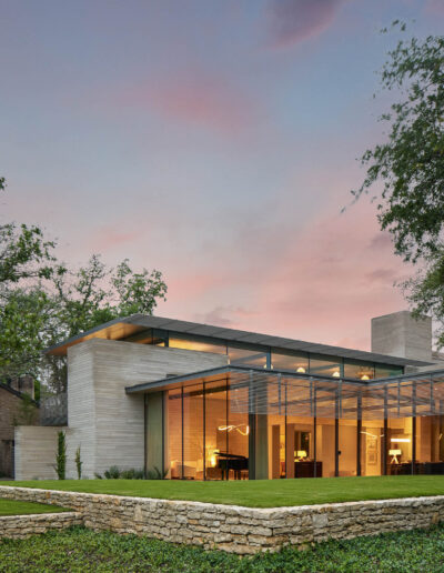 Modern house with large glass windows and a flat roof, surrounded by trees and a manicured lawn, under a sunset sky.
