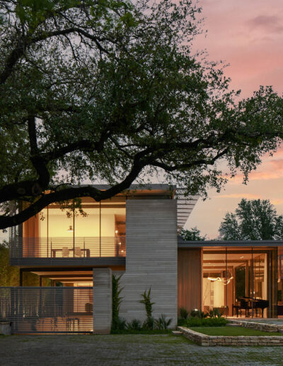 Modern two-story house with large glass windows, surrounded by trees, at sunset. The architecture features clean lines and a mix of wood and concrete elements.