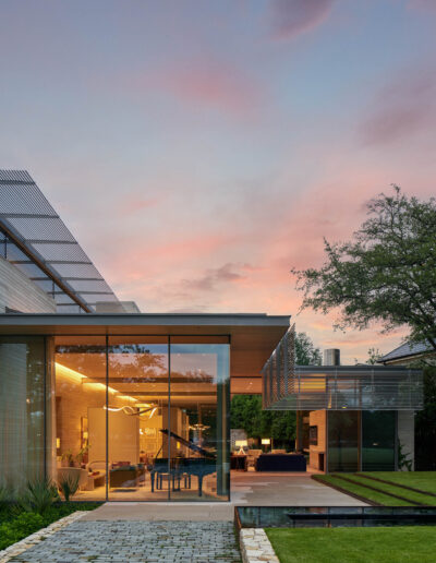 Modern glass house with illuminated interior, surrounded by trees and lawn, features a cobblestone pathway at sunset.