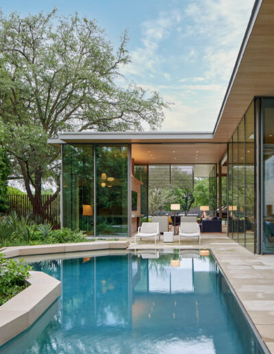 Modern house with floor-to-ceiling glass walls opening to a patio and swimming pool, surrounded by greenery. Outdoor furniture and trees provide a serene setting.