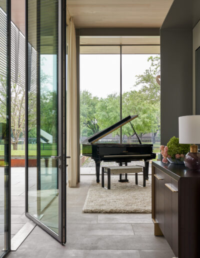 Room with a large window and open glass door, featuring a grand piano on a rug, surrounded by trees outside. A side table with decorative items is nearby.