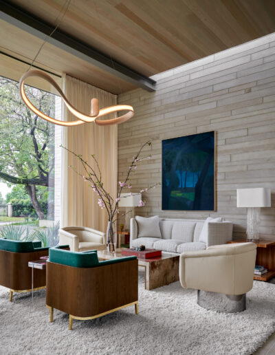 Modern living room with a large loop chandelier, white sofa, two armchairs, and a wooden coffee table. A painting hangs on the stone wall, and large windows show an outside view.