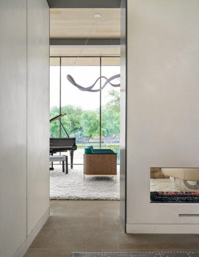 A modern interior with a grand piano, a green armchair, and a fireplace. Large windows overlook a garden. A wavy light fixture hangs from the ceiling.