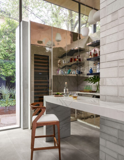 Modern kitchen with high ceiling, reflective cabinets, and a marble countertop. Wooden chair sits by the counter. Large windows reveal outdoor greenery.