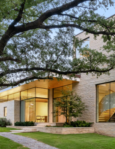 Modern two-story house with large windows and light stone exterior, surrounded by trees and a well-kept lawn, with a visible interior staircase.