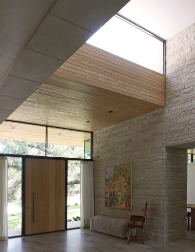 Modern entryway with large wooden door, floor-to-ceiling windows, brick wall, bench, chair, and a colorful painting; natural light fills the space.