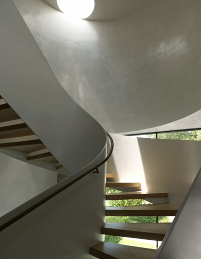 Curved staircase with wooden steps and metal railing, illuminated by natural light from a circular skylight and a window.