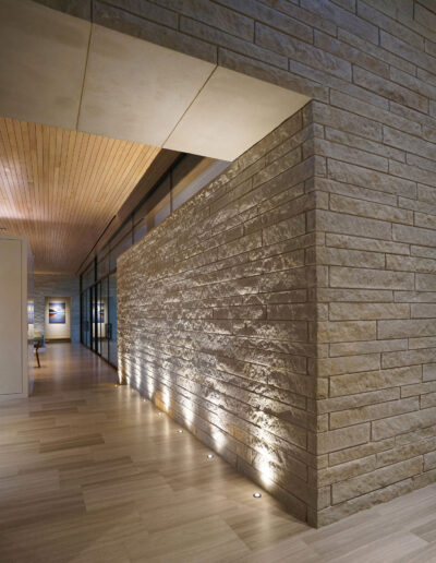 Modern hallway with stone accent wall illuminated by recessed floor lights, wooden ceiling, and framed artwork visible in the background.