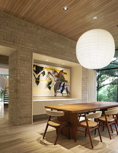 Modern dining room with a wooden table, six chairs, large paper lantern light, abstract wall art, stone walls, and floor-to-ceiling windows overlooking trees.