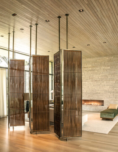 A modern living space features three vertical wooden slatted room dividers, light wood flooring, stone accent wall, linear fireplace, large windows, and a ceiling with recessed lights.