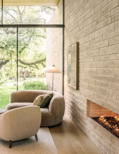 Modern living room with beige furniture, a round coffee table, a built-in fireplace, large floor-to-ceiling windows, and a view of green trees and grass outside.