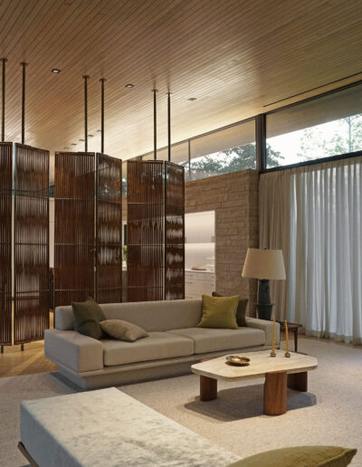 Modern living room with neutral-toned furniture, large windows, wooden ceiling, partition shelves, and natural light streaming in from outside.