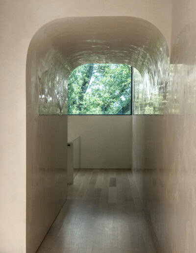 A smooth, curved hallway with light-colored walls leads to a large window overlooking green trees outside.