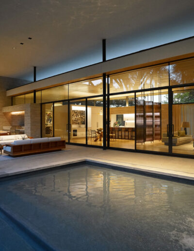 Modern house with large glass walls opens to a patio with a small pool and outdoor seating area, revealing a well-lit, stylish interior living and dining space.