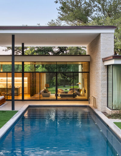 Modern house with large glass windows and a swimming pool in the foreground, surrounded by greenery and trees.