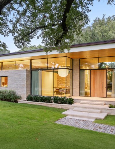 Modern house with large glass windows, stone exterior, and a well-manicured lawn, surrounded by trees and greenery.