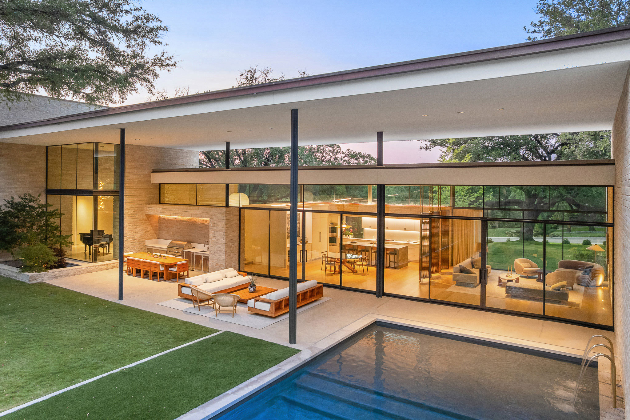 Modern house with large glass windows, outdoor seating area, and swimming pool overlooking a grassy yard at sunset.